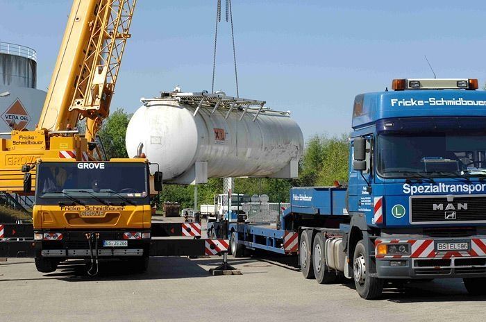 Ein Kran hebt einen großen Tank auf einen Schwertransporter auf einem Industriegelände.